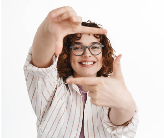 A young woman with curly hair, wearing glasses and a striped shirt, smiles as she uses her hands to frame a scene, suggesting